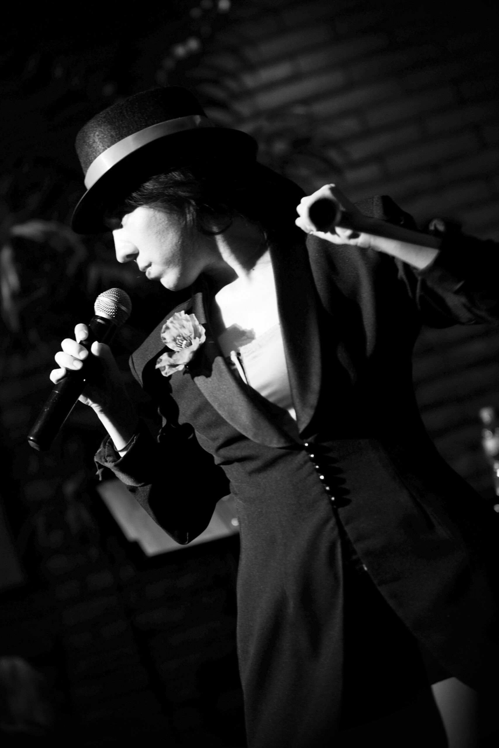 Black and white photo of a woman singing into a microphone, wearing a hat and blazer.