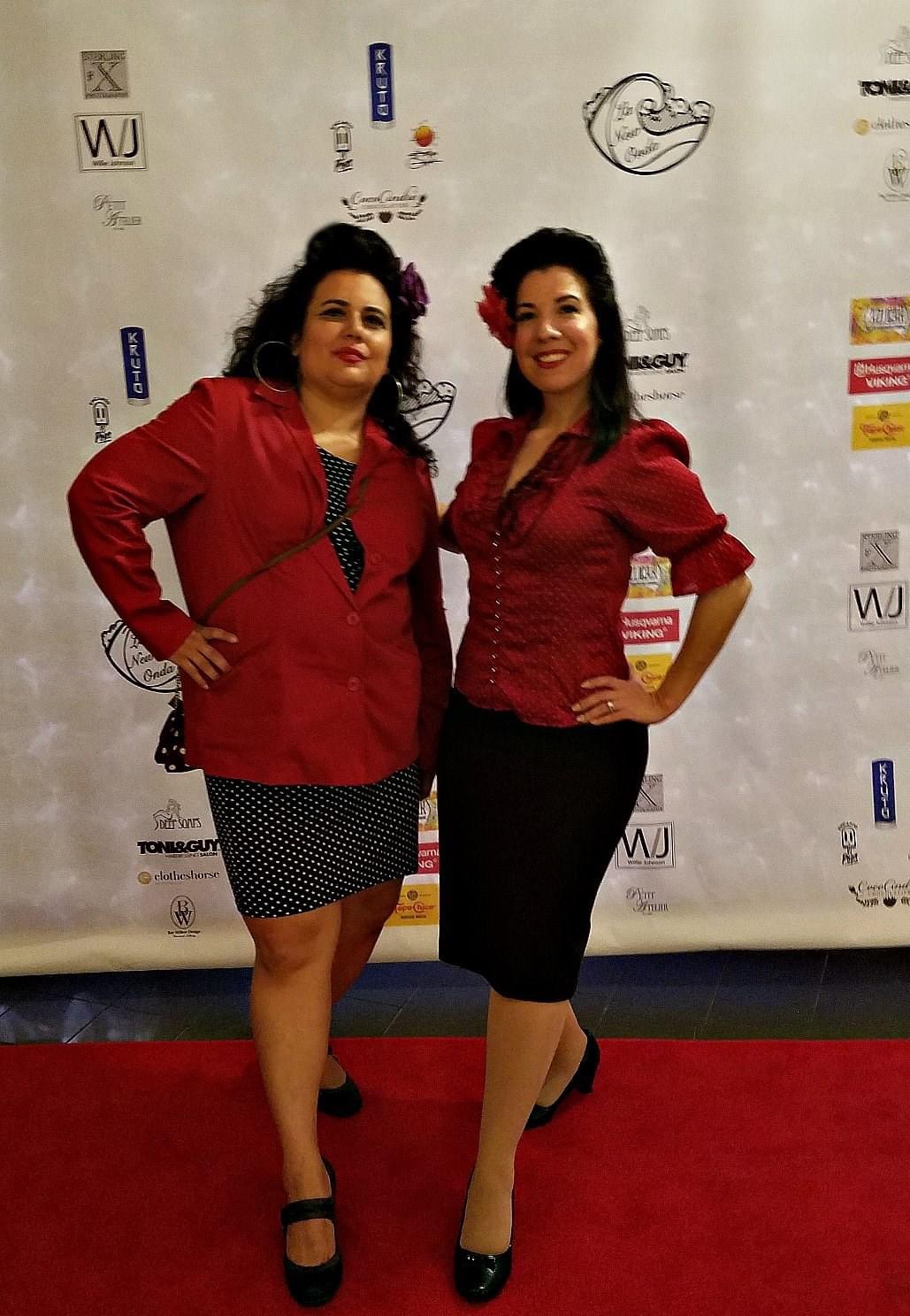 Two women posing confidently on a red carpet in red tops and skirts.