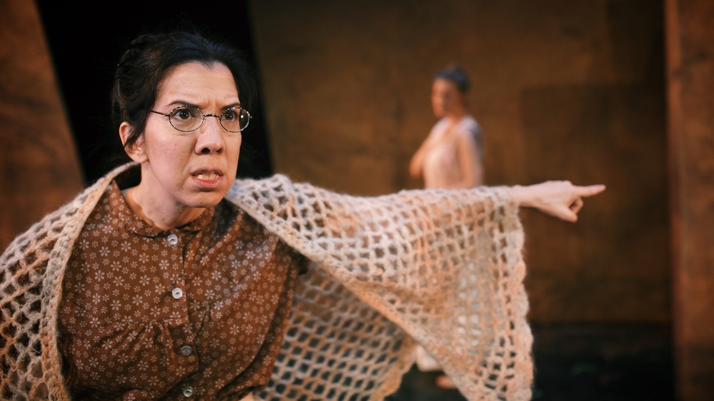 A tense woman in glasses holds a crocheted shawl, with a blurred figure behind her.