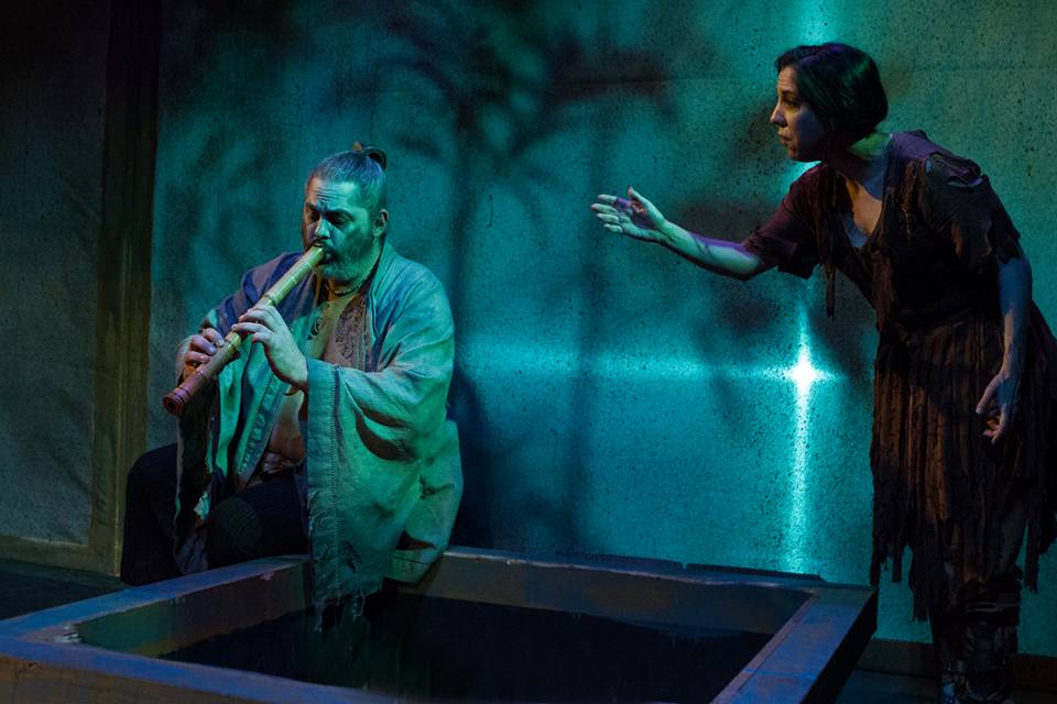 A man playing a flute in a boat with a woman reaching out to him under blue lighting.