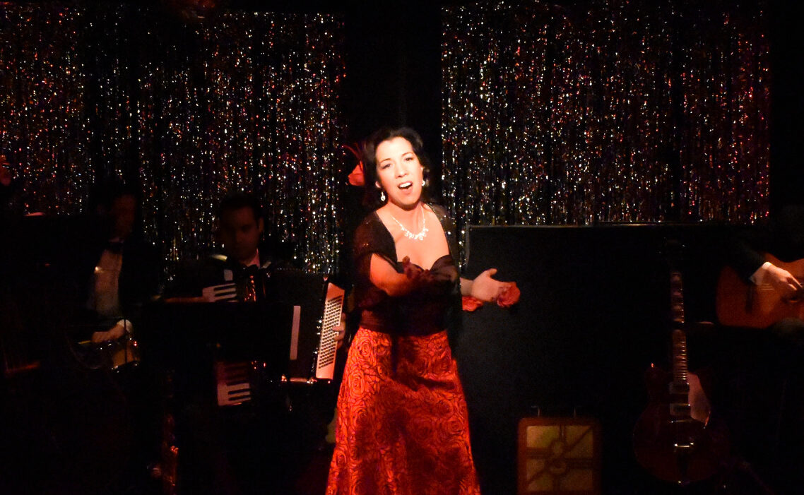 A woman passionately singing on stage in a red dress with dark background.