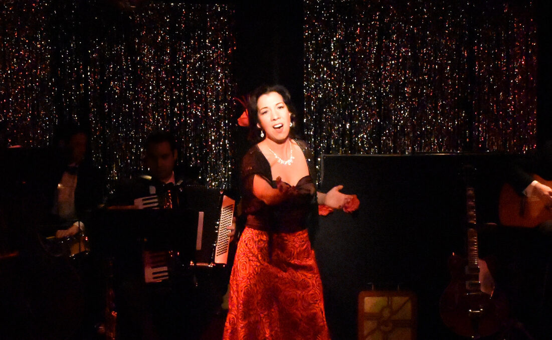A woman passionately singing on stage in a red dress with a pianist in the background.