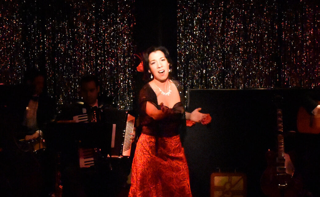A passionate woman sings on stage in a red dress with an accordion player behind her.