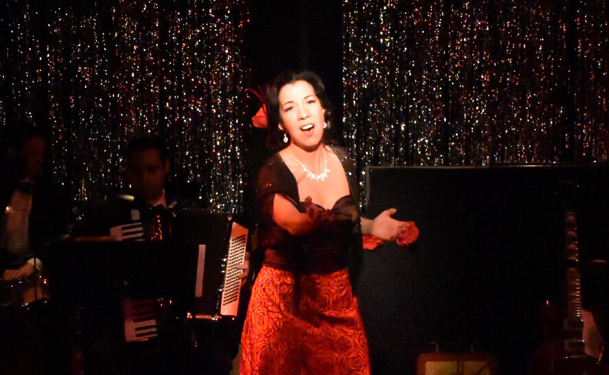 A woman passionately singing on stage in a red dress with a sparkling backdrop.