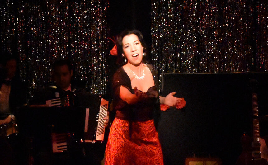A woman passionately singing on stage in a red dress with a sparkling backdrop.