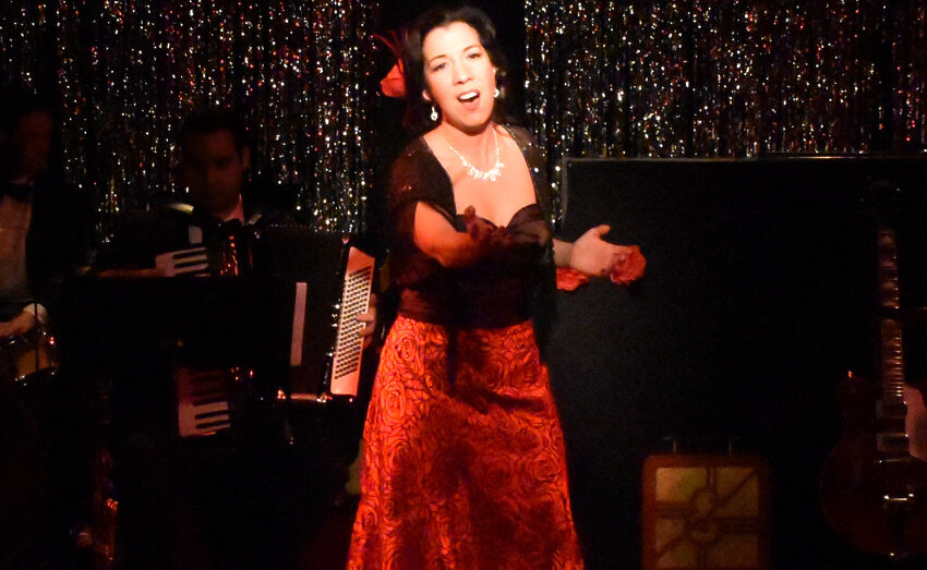 A woman passionately singing on stage in a red dress with an accordion player behind her.