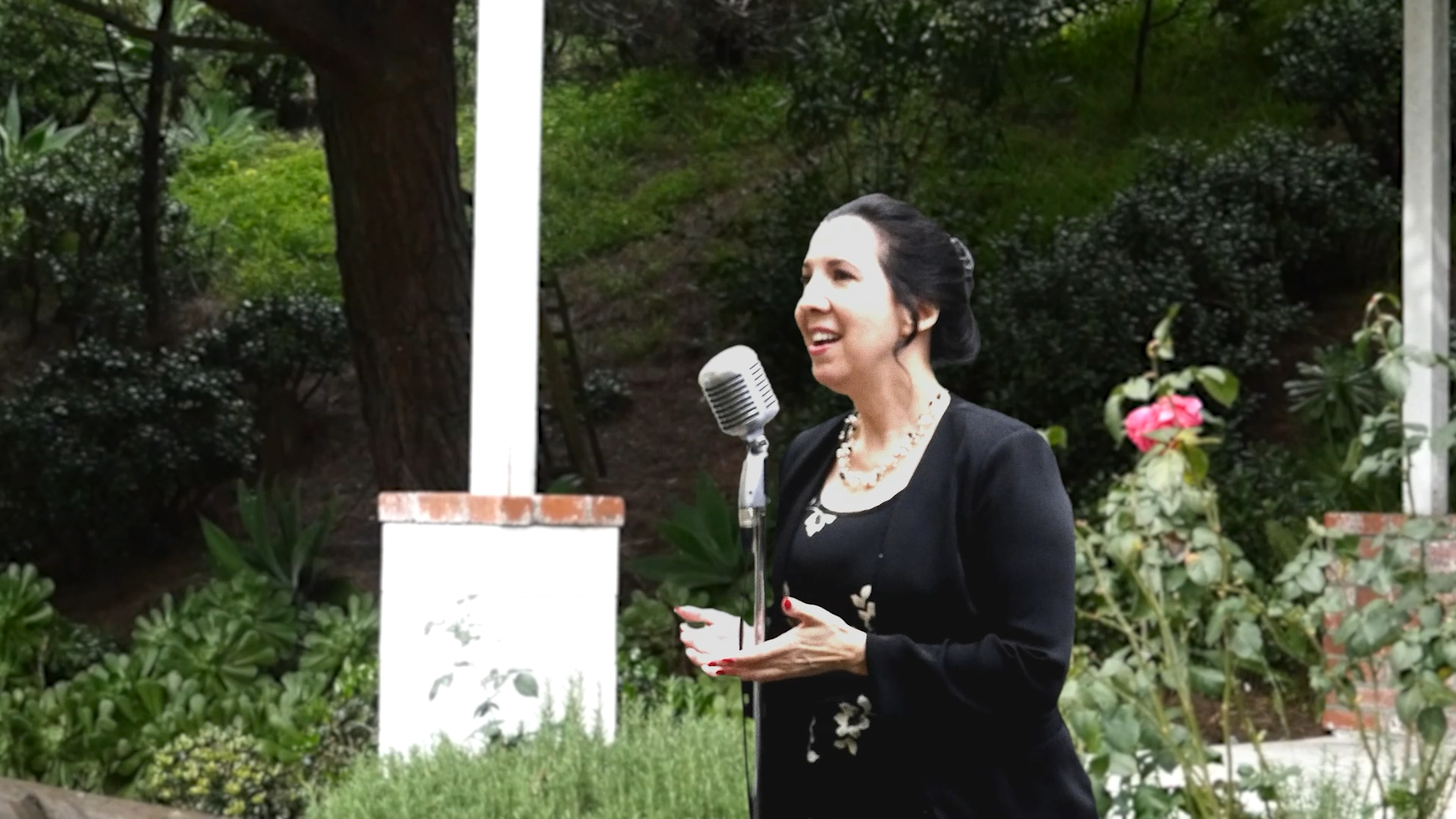 A woman speaks into a vintage microphone outdoors near a tree.
